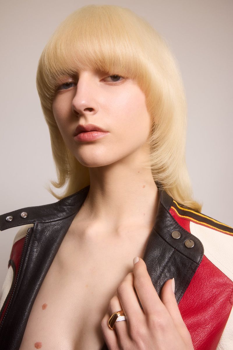 Close-up portrait of a young adult female with blonde, fringed hair wearing a leather jacket against a neutral background.