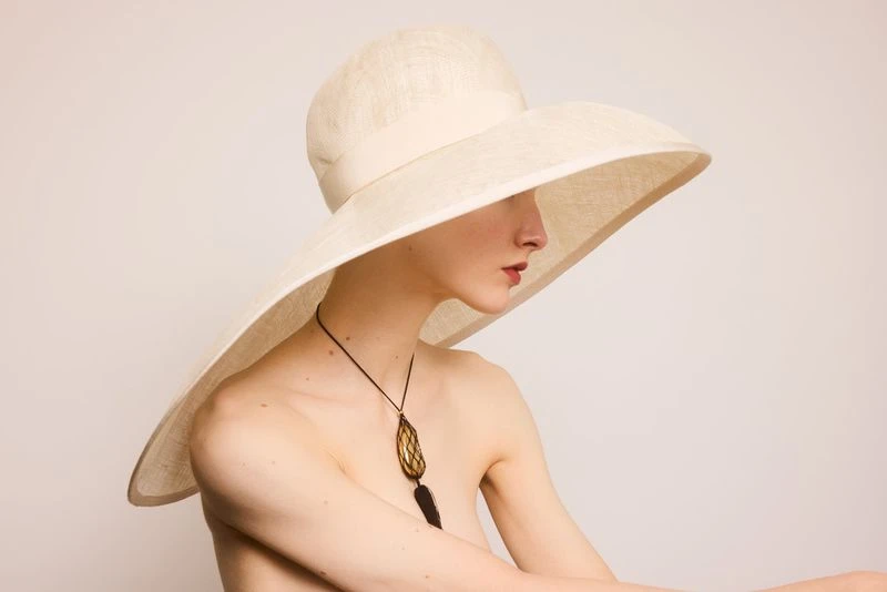 Woman with bare shoulders poses in a cream-colored wide-brimmed hat and necklace against a neutral background in a beauty shot.