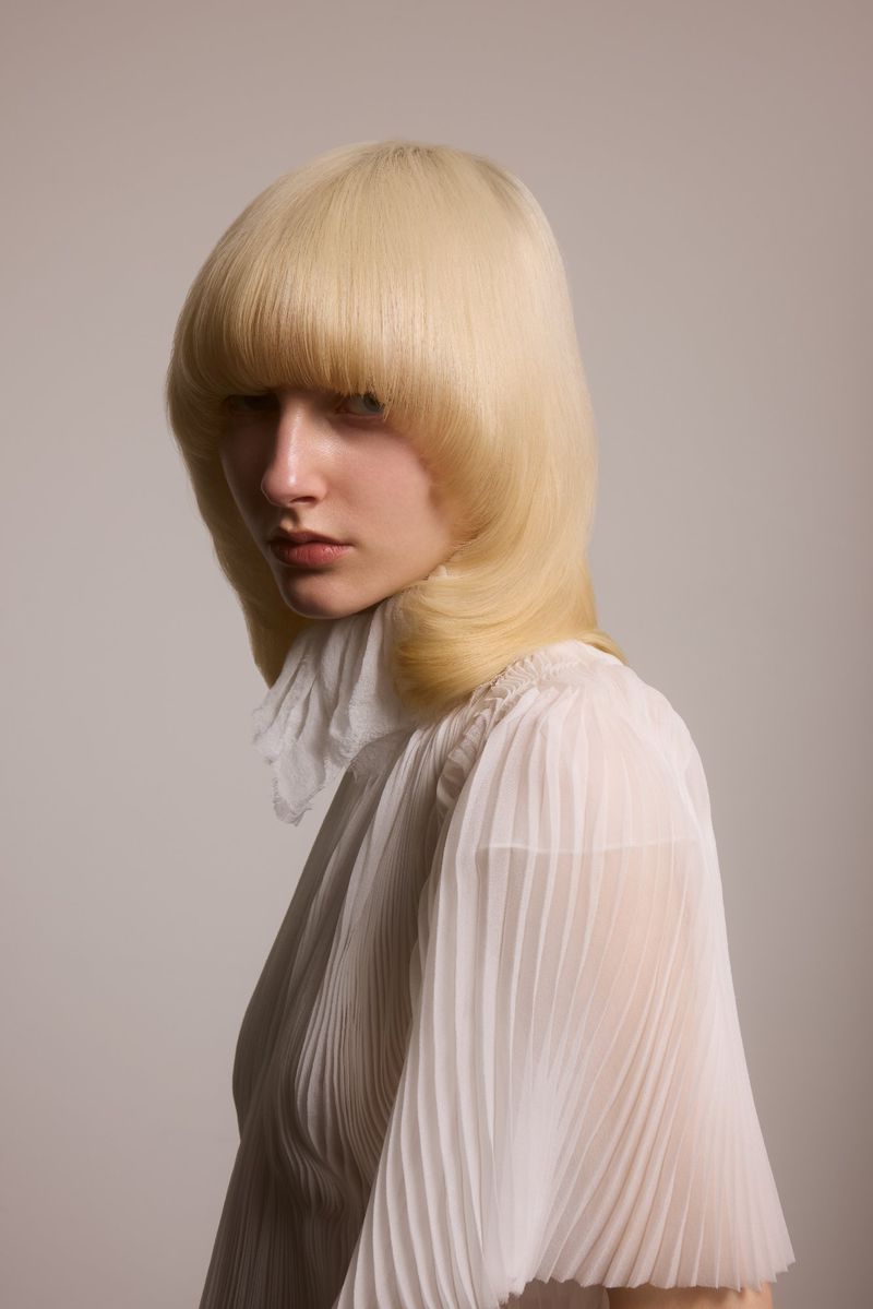 Young woman with a blonde bowl haircut and pale skin poses in front of a neutral backdrop, wearing a translucent white blouse.
