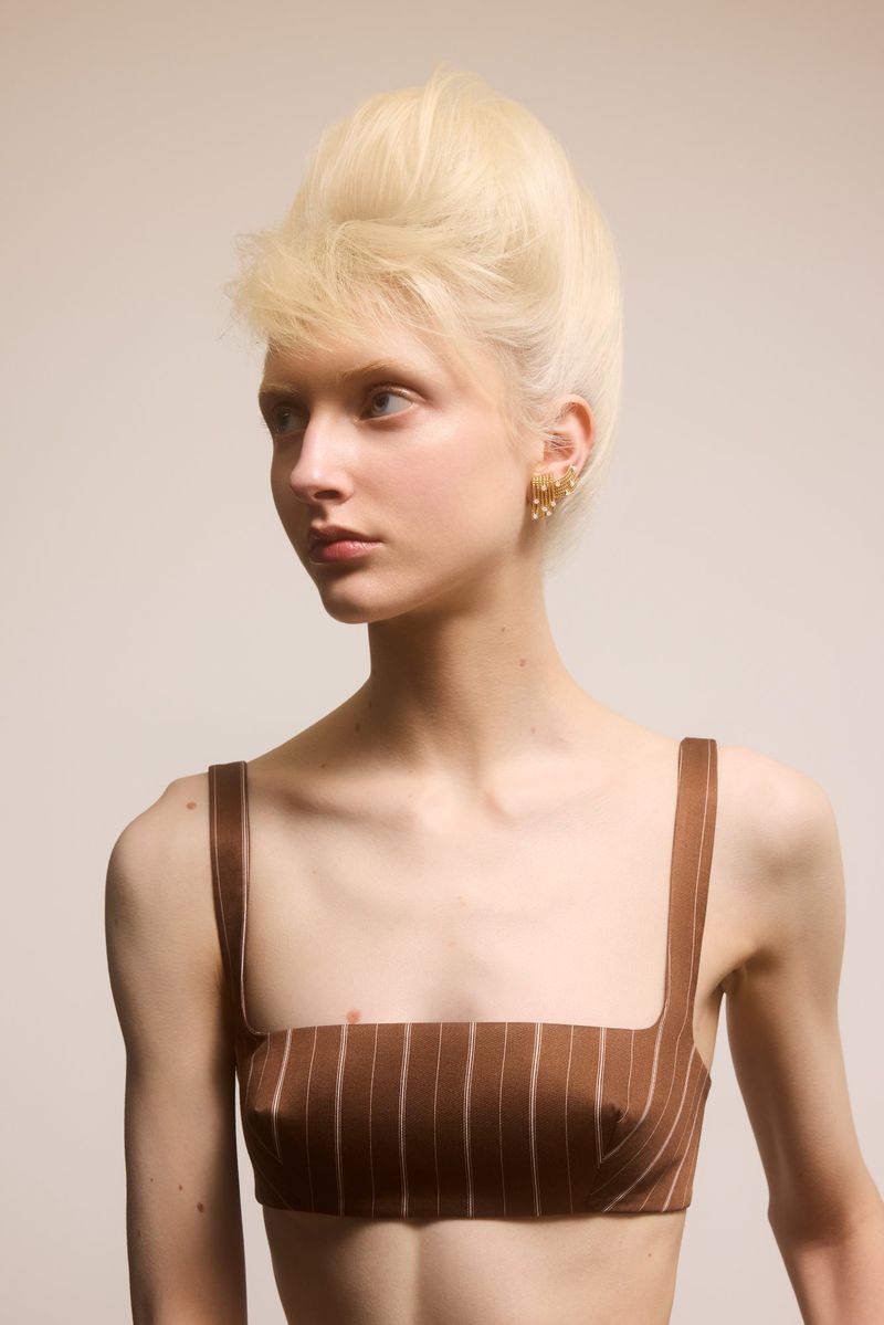 Portrait of a young woman with a striking blonde beehive hairstyle, wearing a brown pinstripe top, gazing off-camera in a neutral studio setting.