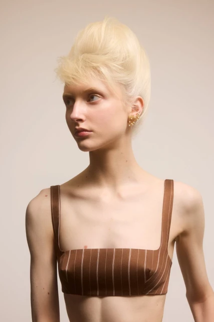 Portrait of a young woman with a striking blonde beehive hairstyle, wearing a brown pinstripe top, gazing off-camera in a neutral studio setting.