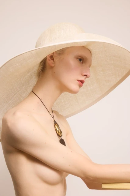 Blonde woman poses wearing large sun hat and necklace in a close-up portrait with soft lighting on a seamless backdrop.