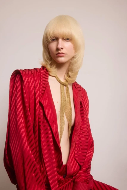 Adult female in red suit with gold necklace and blonde hair poses in front of neutral background in a fashion studio setting.
