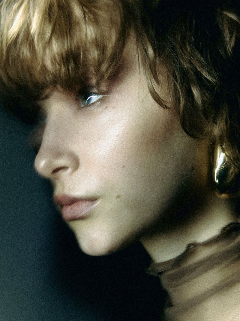 Close-up studio portrait of a young woman with short brown hair and a neutral facial expression wearing a brown top. Fashion, beauty.