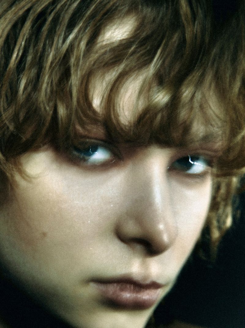 Close-up portrait of a young adult with fair skin, light brown wavy hair, and blue eyes looking directly at the camera in soft, diffused lighting.