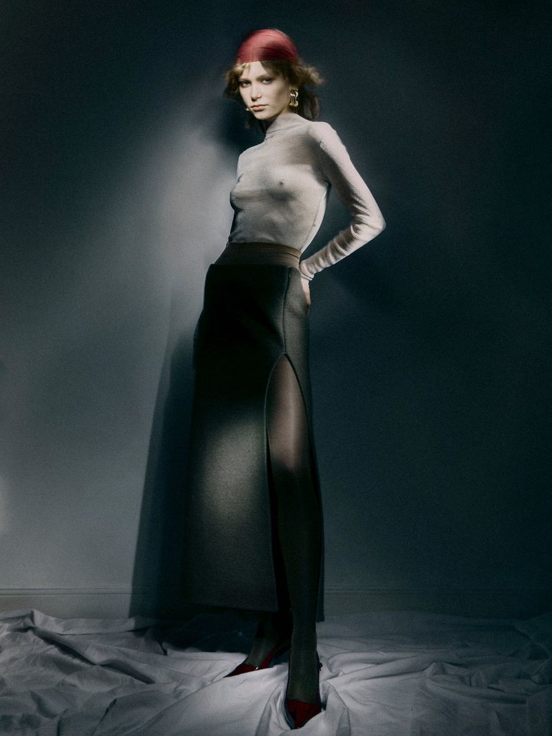 A stylish adult female stands with hands on hips, wearing a long-sleeved top, skirt with slit, red heels, and head covering in a studio setting.
