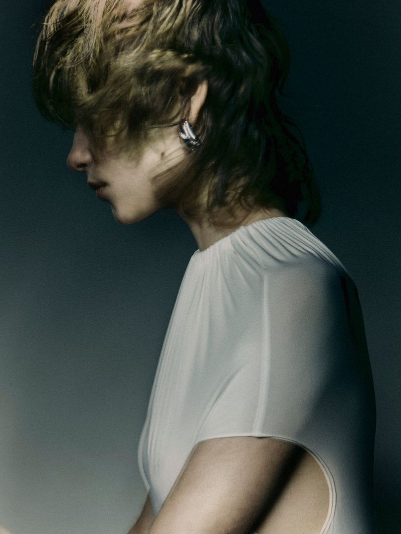 Profile shot of a fashion model with short shaggy hair, pale complexion, wearing an off-white top with shoulder cut-outs against a dark background.