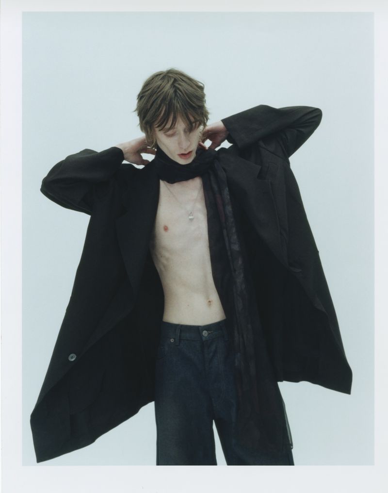 Model Casper Tyborn wearing an open black coat and dark trousers against a plain light studio background for The Greatest Magazine.