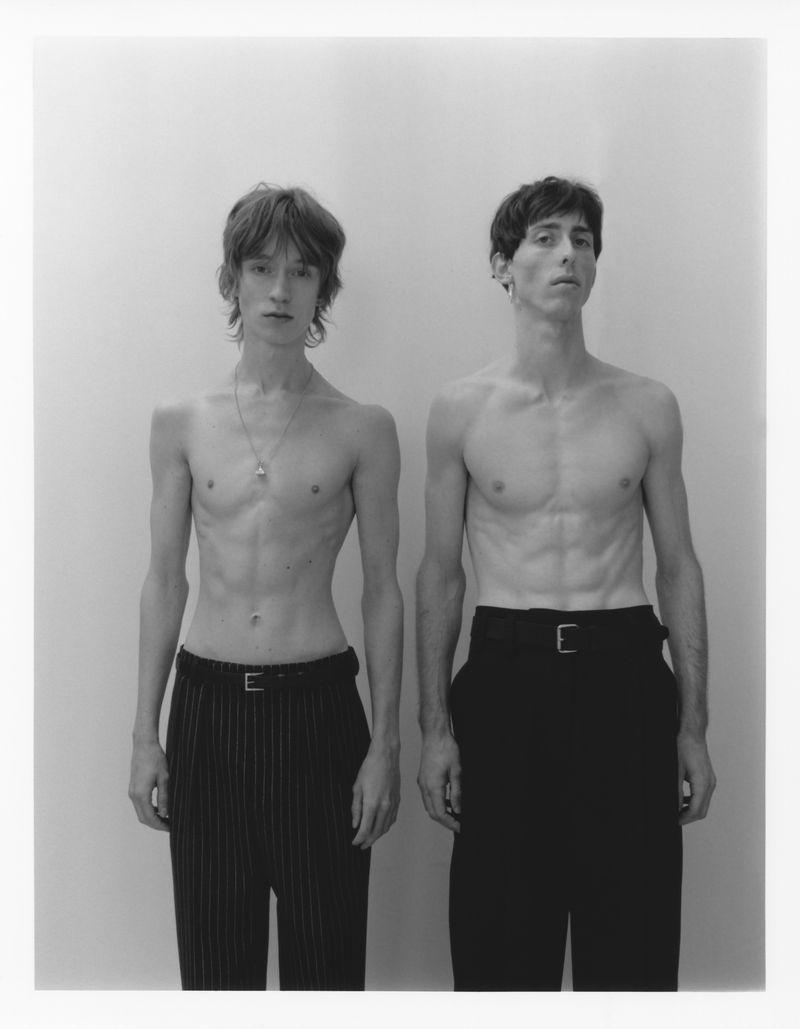 Black and white portrait of two shirtless adult males with dark hair, wearing black pants with belts, posing in a photo studio.