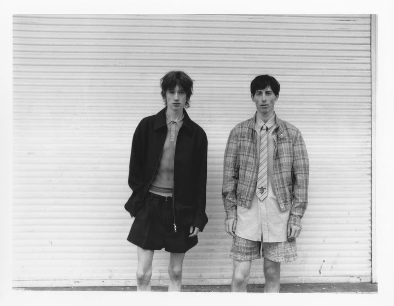 Black and white shot of two slender male fashion models in jackets and shorts standing against a corrugated metal wall.