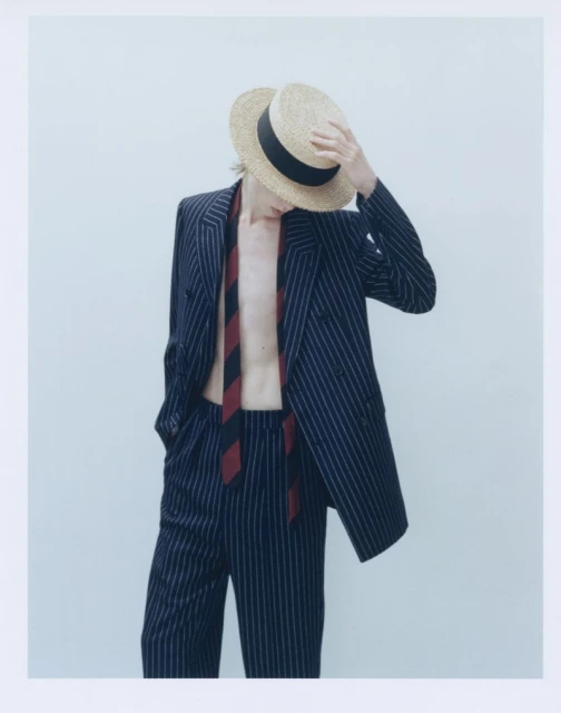 Fashionable person in a pinstripe suit and boater hat poses against a white background in a studio setting.