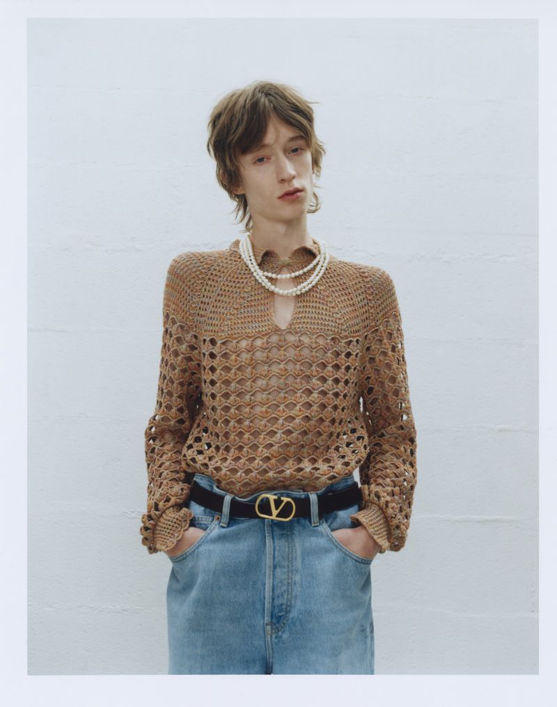 A person in a tan, long-sleeved, crocheted shirt, black belt, and light blue jeans poses in front of a solid white background.