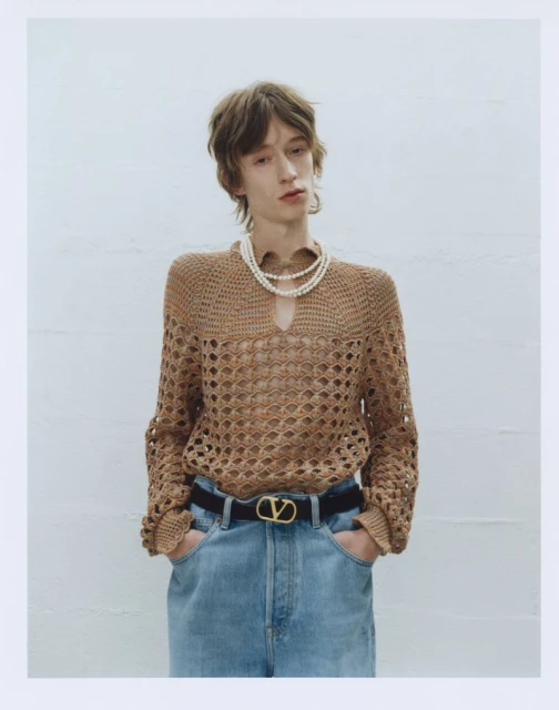 A person in a tan, long-sleeved, crocheted shirt, black belt, and light blue jeans poses in front of a solid white background.