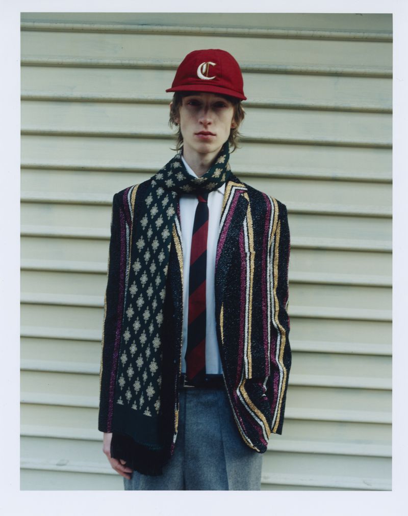 Fashionable adult male poses in front of a light-colored wall, wearing a striped blazer, scarf, tie, cap, and grey pants in an outdoor setting.