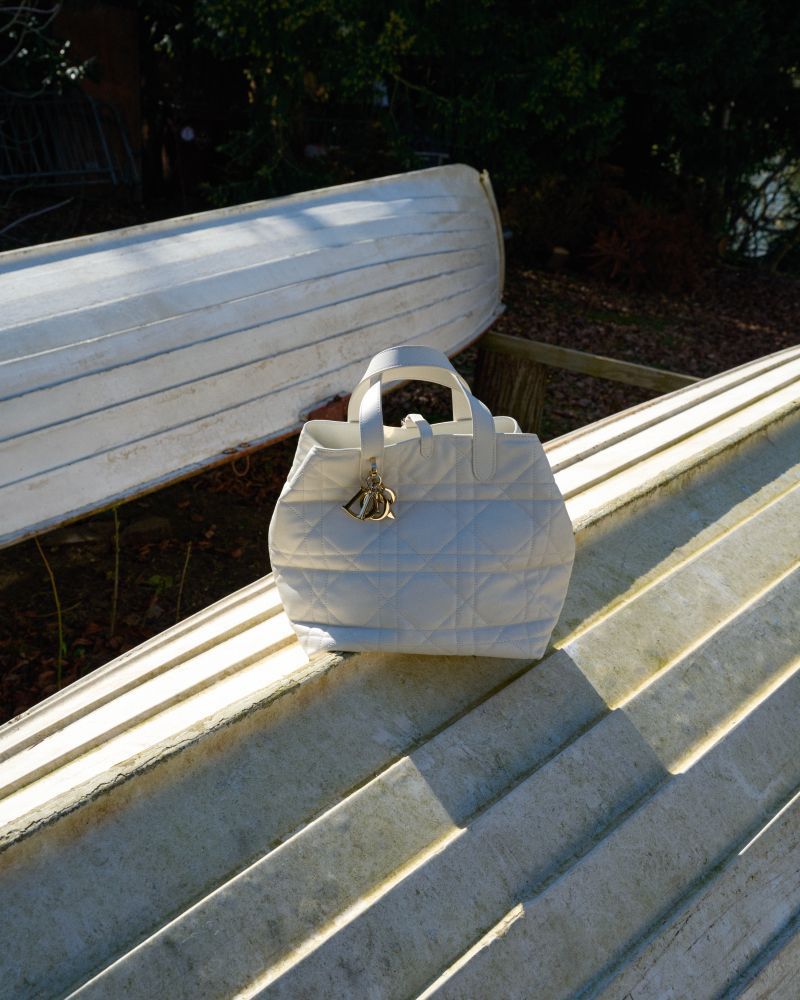 White quilted tote handbag with a decorative charm, sitting on a series of steps outdoors with green trees in the background.