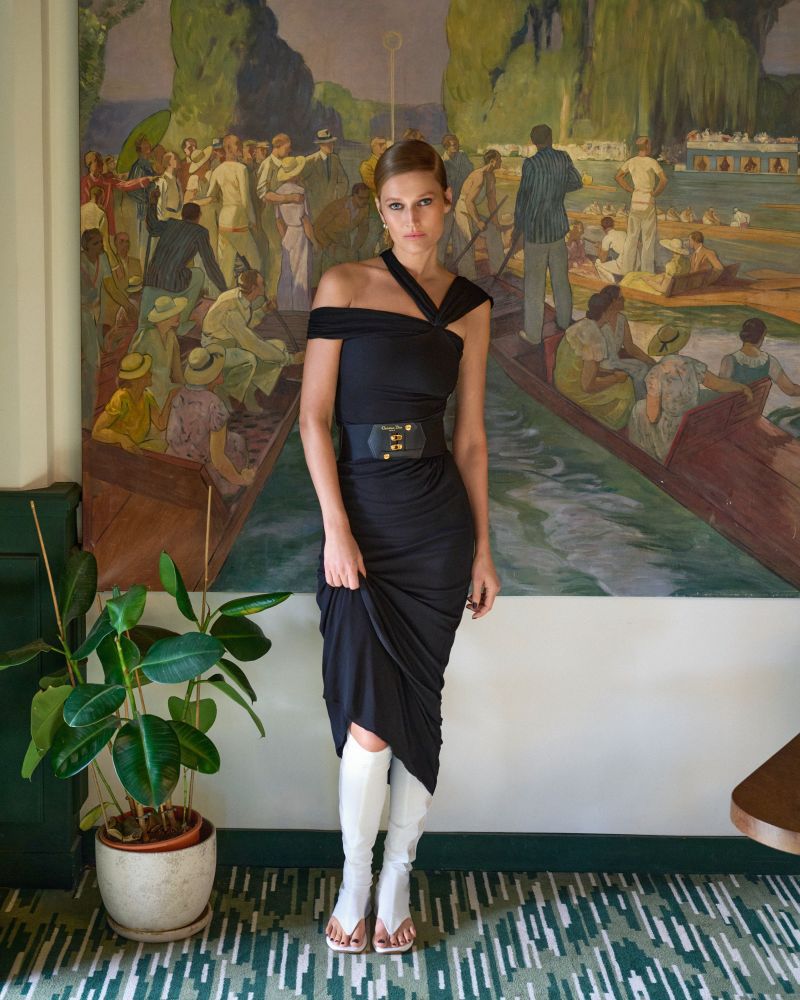 Elegant female fashion model posing in a black dress with a wide belt and white high boots in front of a vintage wall art.
