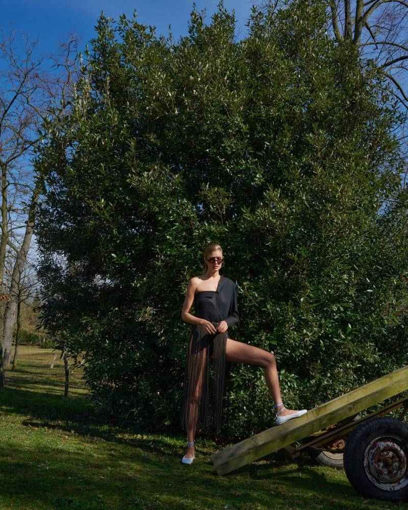 An adult female poses in a one-shoulder black dress and see-through skirt in a grassy outdoor setting with large tree backdrop.