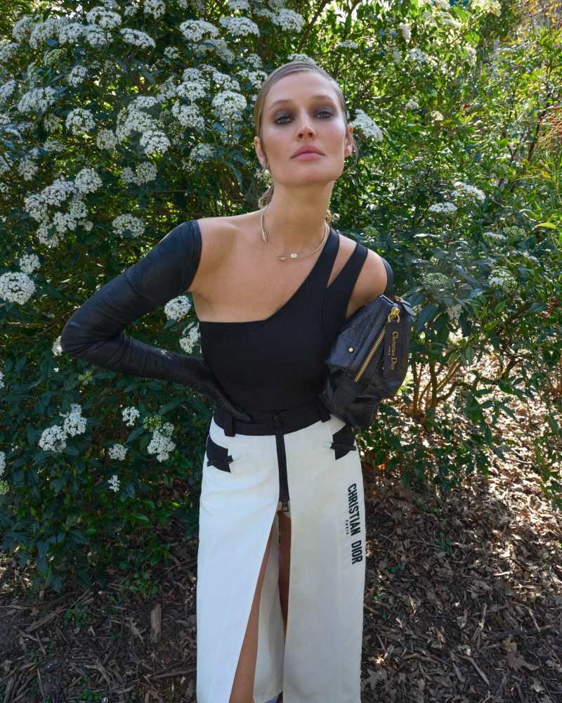A woman poses outdoors wearing a black one-shoulder top, cream skirt with black text, black gloves, and a black purse against a bush.