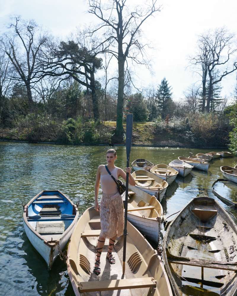 A woman stands among rowboats on a lake in a fashionable skirt and sandal heels holding an oar on a sunny day.