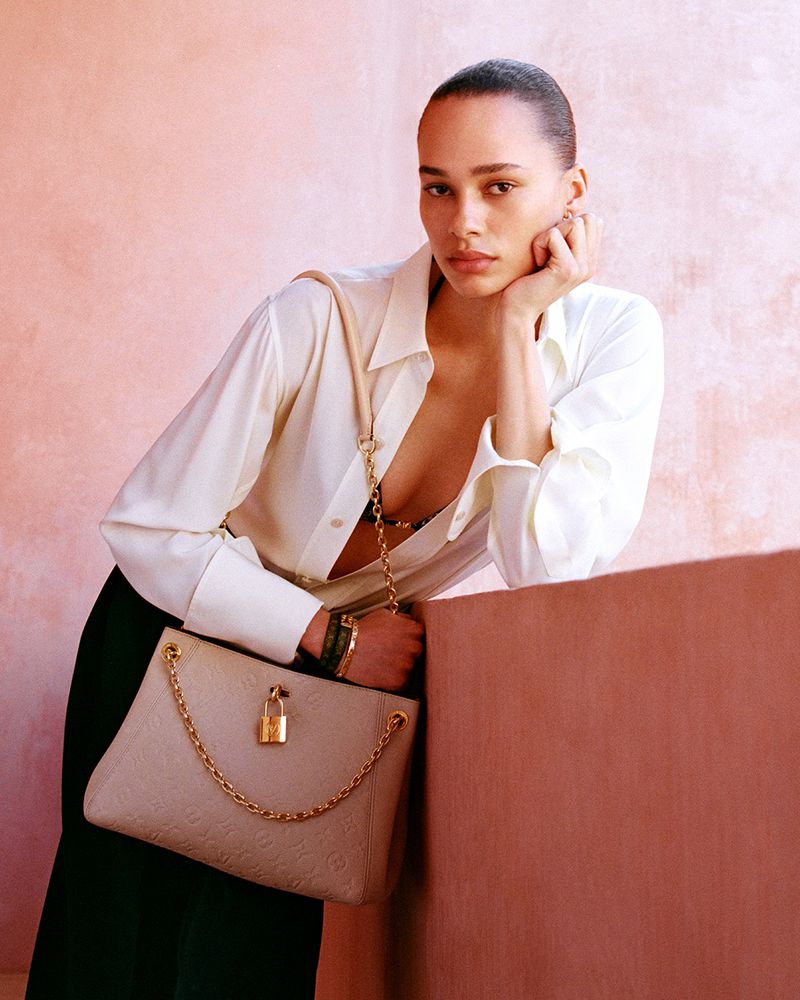 Model leaning on a ledge wearing a white shirt and holding a beige shoulder bag for Louis Vuitton