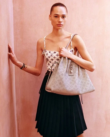 Model standing against a pink wall wearing a monogram top and dark pleated skirt, holding a grey shoulder bag