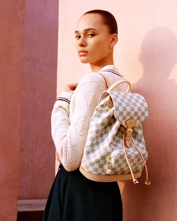 Woman poses with a Louis Vuitton Damier Azur backpack in front of a textured pink wall, wearing a white knit sweater and dark skirt.