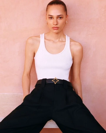 Model Annemary Aderibigbe in a white ribbed tank top and black pants with a gold-accented belt against a pink wall