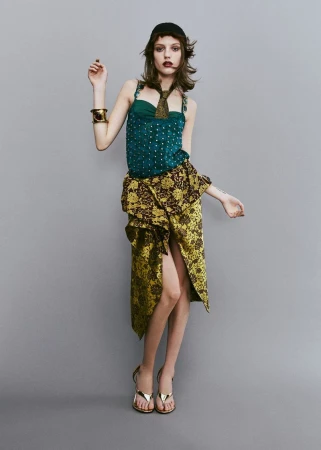 Model wearing a patterned green top and gold patterned skirt against a neutral grey studio background