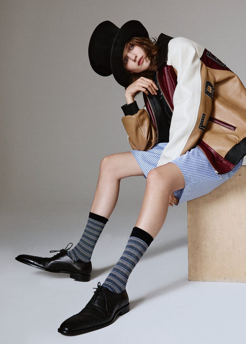 A woman in a black hat and tan varsity jacket sits in a studio, modeling striped shorts and socks with black leather dress shoes.