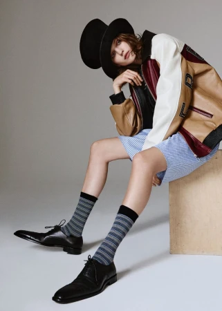 A woman in a black hat and tan varsity jacket sits in a studio, modeling striped shorts and socks with black leather dress shoes.