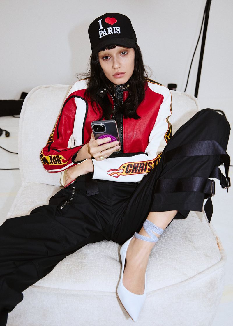 Woman poses in a racing-style jacket and black pants, seated on white chair, holding smartphone with an 'I Heart Paris' cap.