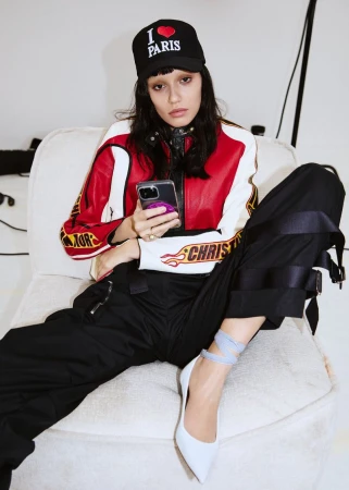 Woman poses in a racing-style jacket and black pants, seated on white chair, holding smartphone with an 'I Heart Paris' cap.