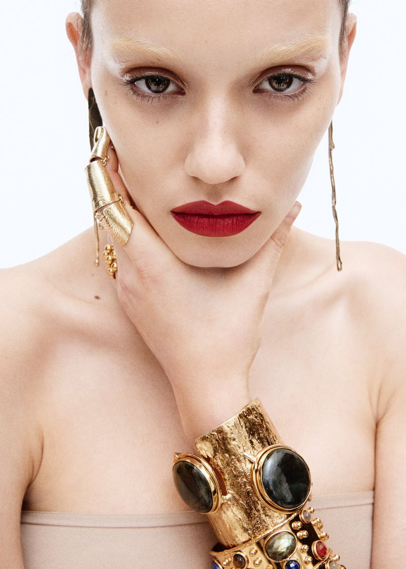 Close-up portrait of a woman wearing gold statement jewelry and dark red lipstick, set against a bright white background.
