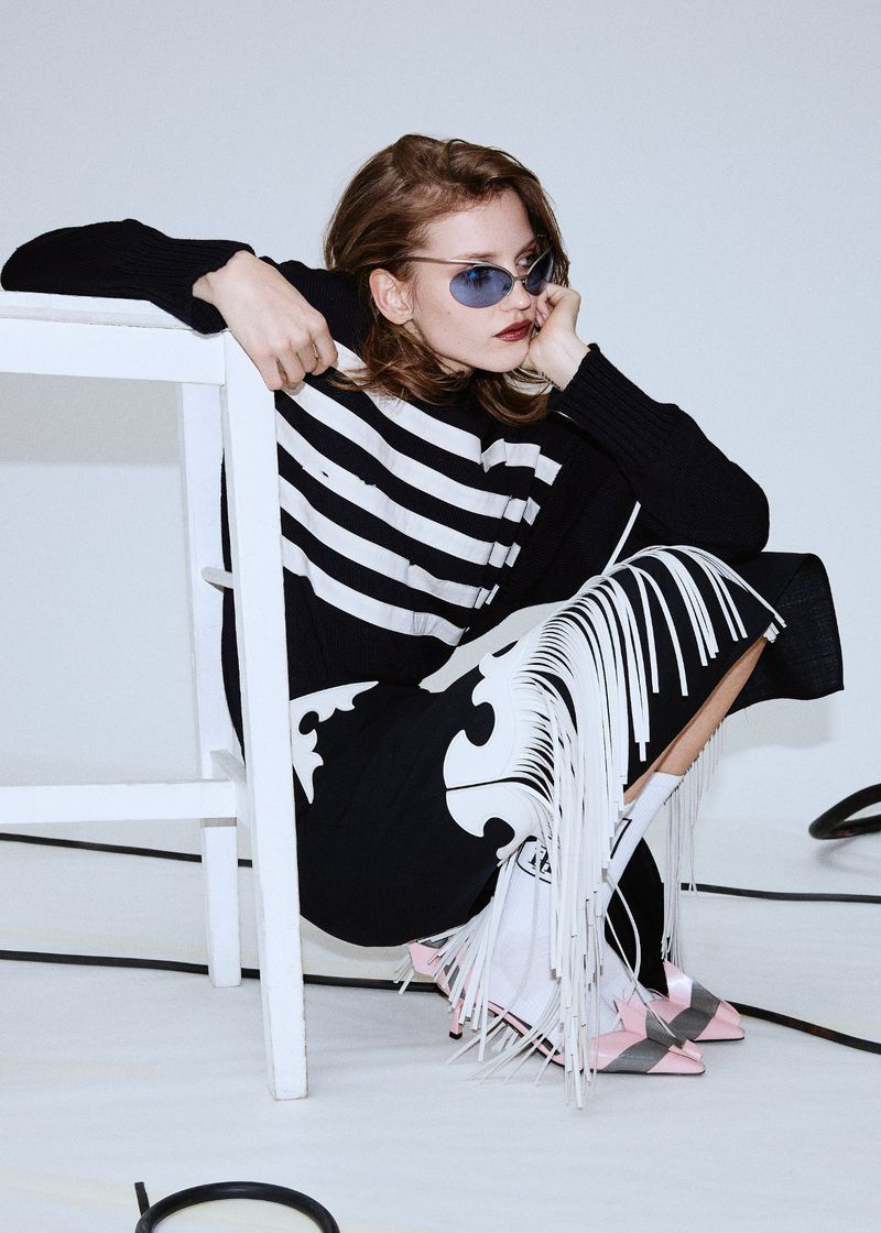 A stylish woman wearing sunglasses, a black and white striped sweater, and a fringed skirt sits on a white stool in a bright studio.