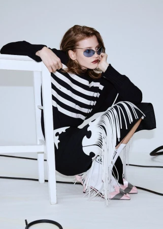 A stylish woman wearing sunglasses, a black and white striped sweater, and a fringed skirt sits on a white stool in a bright studio.