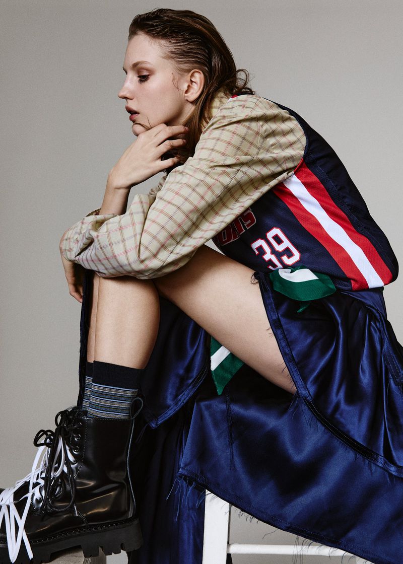 Young woman in a sports-inspired ensemble poses indoors on a stool, showcasing fashion, style, and athletic wear in a studio setting.