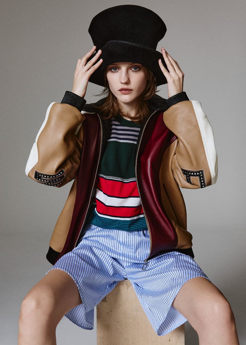 A young woman wearing a large black hat and a colorful jacket poses while sitting on a wooden block in a photo studio setting.