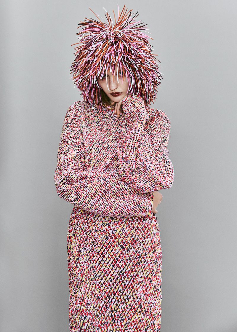 Woman poses in studio wearing a colorful, textured sweater and skirt with a matching wig. Studio lighting and gray background.