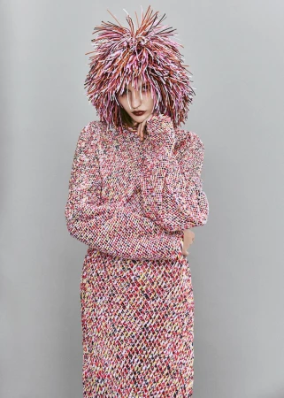 Woman poses in studio wearing a colorful, textured sweater and skirt with a matching wig. Studio lighting and gray background.