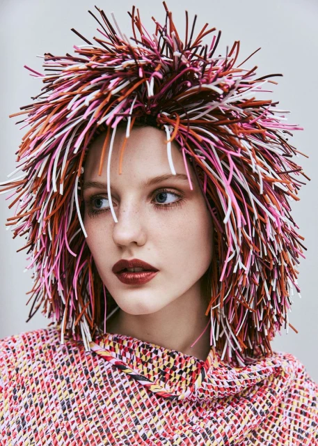 Portrait of a female model wearing a multicoloured woven top and a vibrant, spiky headpiece with white background in studio lighting.