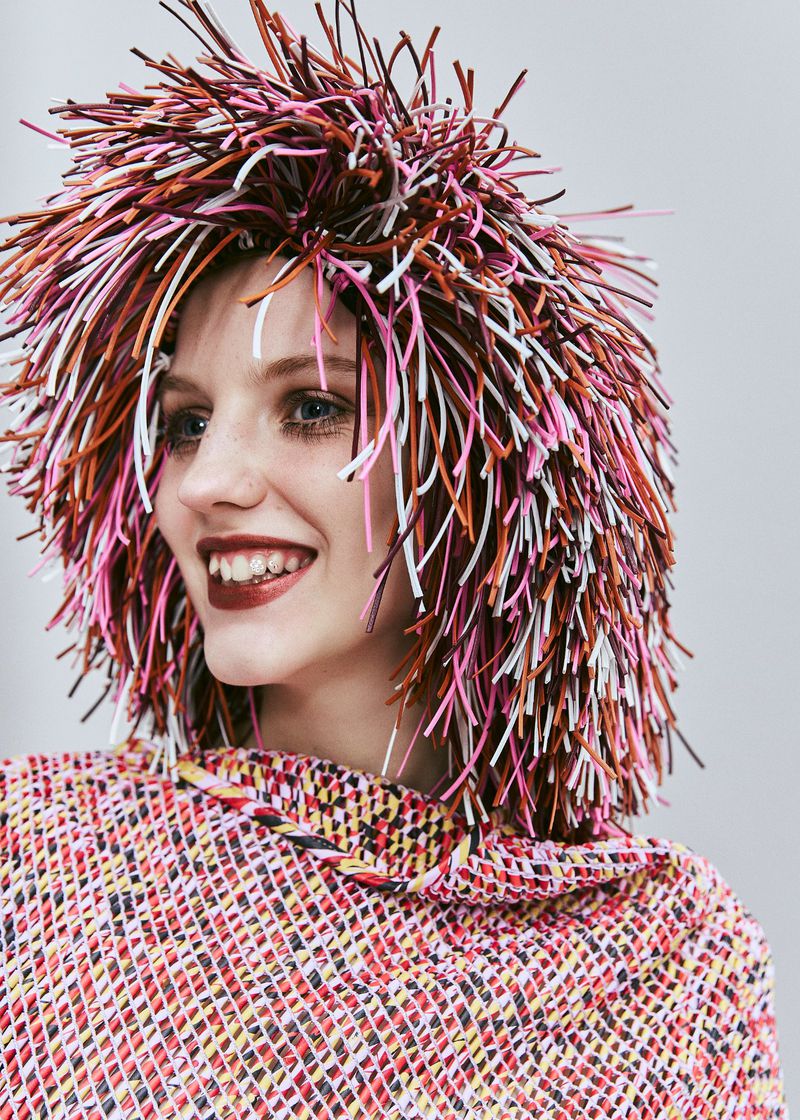 A smiling woman with a vibrant, multicolored wig and a matching top, set against a light gray background, radiates a playful and artistic vibe.