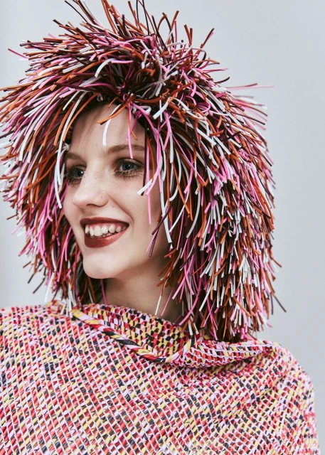A smiling woman with a vibrant, multicolored wig and a matching top, set against a light gray background, radiates a playful and artistic vibe.