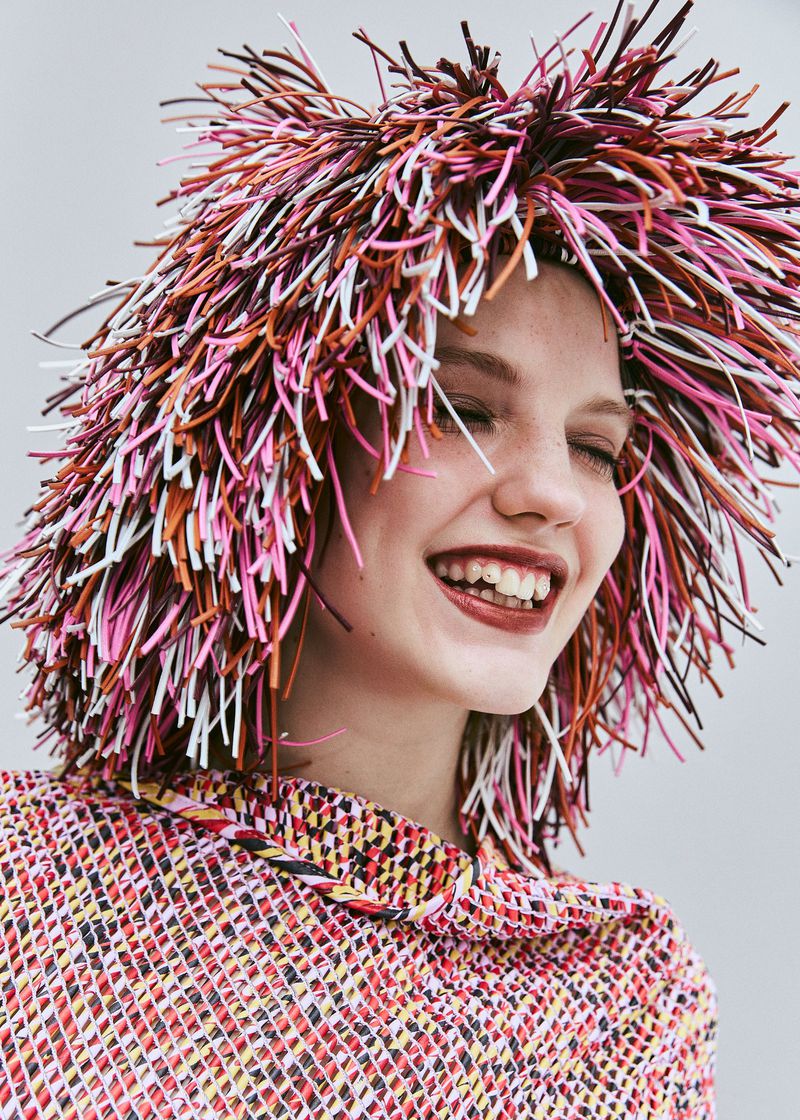 A smiling woman with fair skin wears a multi-colored hat resembling straw, with a matching top and a plain neutral background.