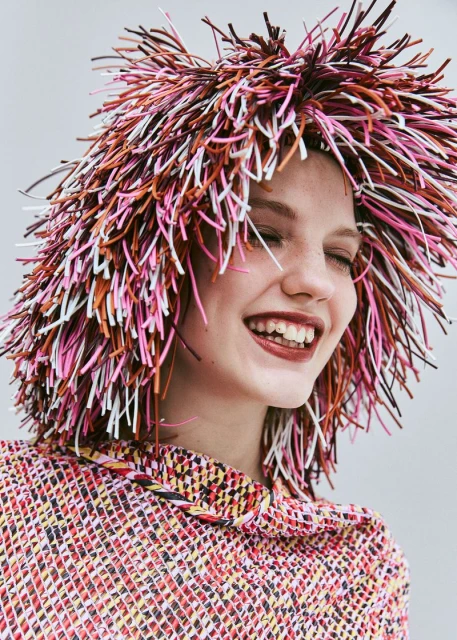 A smiling woman with fair skin wears a multi-colored hat resembling straw, with a matching top and a plain neutral background.