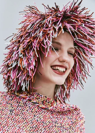 A smiling woman with fair skin wears a multi-colored hat resembling straw, with a matching top and a plain neutral background.