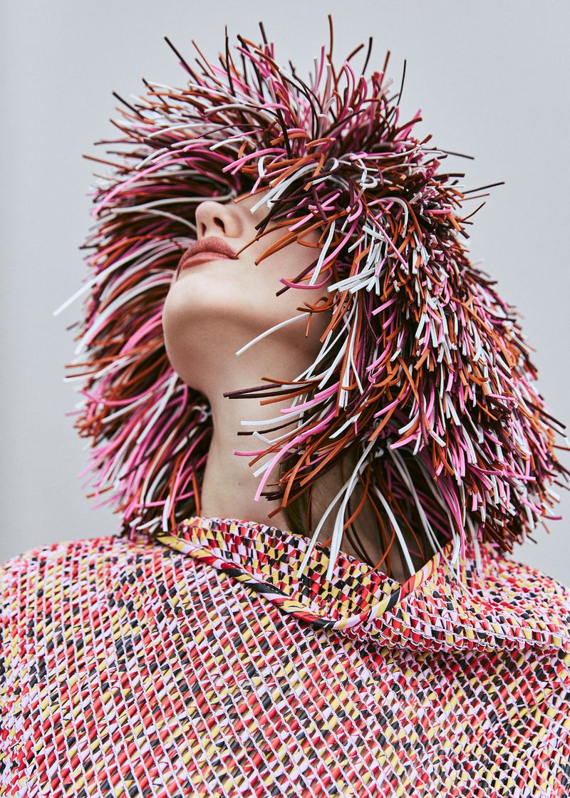 Woman in colorful textured top and vibrant spikey headpiece, looking up in a studio setting. Beauty and fashion editorial shot.