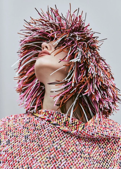 Woman in colorful textured top and vibrant spikey headpiece, looking up in a studio setting. Beauty and fashion editorial shot.