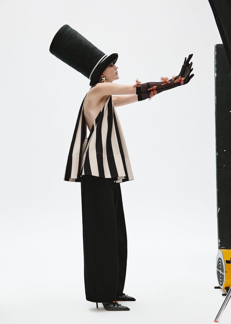 Elegant woman in a striped top and black pants poses with extended arms, wearing a tall black hat and gloves in a bright studio.