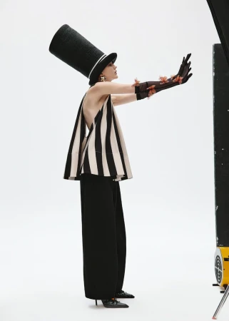 Elegant woman in a striped top and black pants poses with extended arms, wearing a tall black hat and gloves in a bright studio.