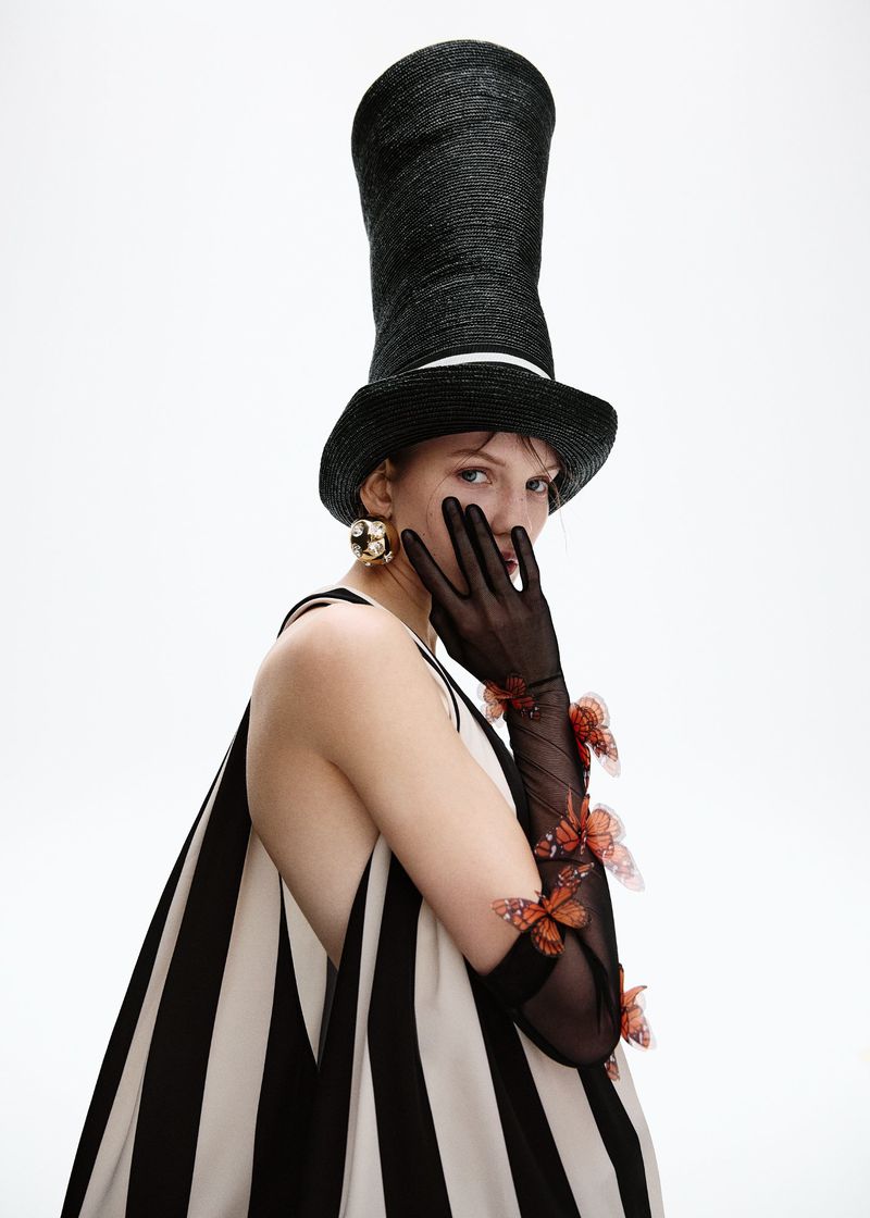 Adult woman poses in a black-and-white striped dress, a black top hat, black gloves with orange butterfly appliques against a white backdrop.
